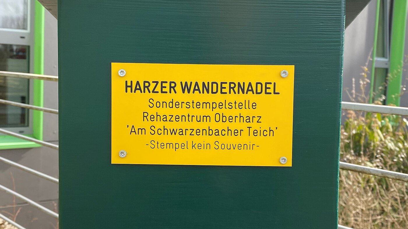 Gelbe Plakette mit schwarzem Text: 'HARZER WANDERNADSEL Sonderstempelstelle Rehazentrum Oberharz 'Am Schwarzenbacher Teich' - Stempel kein Souvenir'.