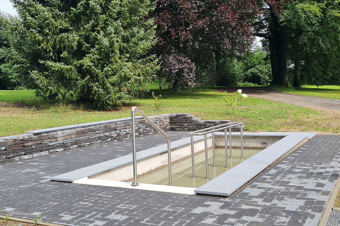Ein Kneipptretbecken mit grauem Steinrand und Edelstahlgeländer. Der Pool ist teilweise mit Wasser gefüllt und befindet sich in einer grünen Umgebung.