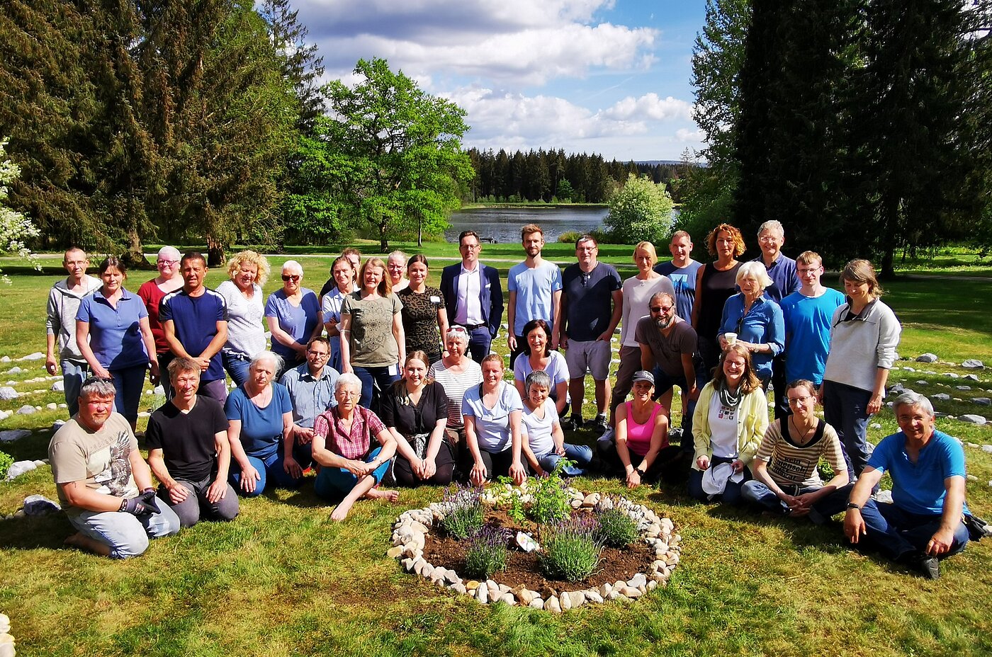 Gruppenfoto von 36 Personen im Freien, umgeben von Bäumen und einem Teich. In der Mitte ein runder Steingarten mit verschiedenen Pflanzen.